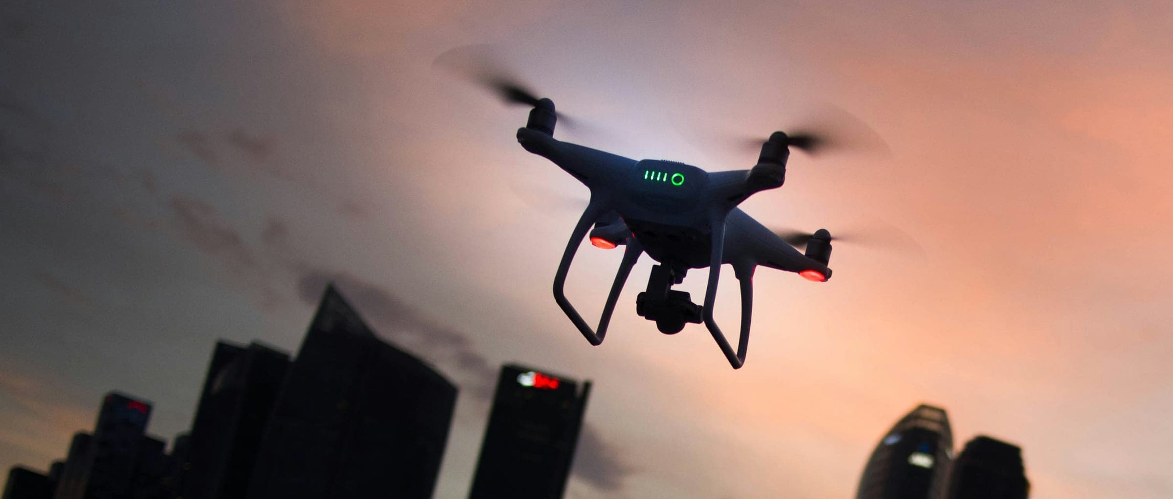 A rotorcraft on station over a city skyline at dusk, its gimbal camera and navigation lights lit