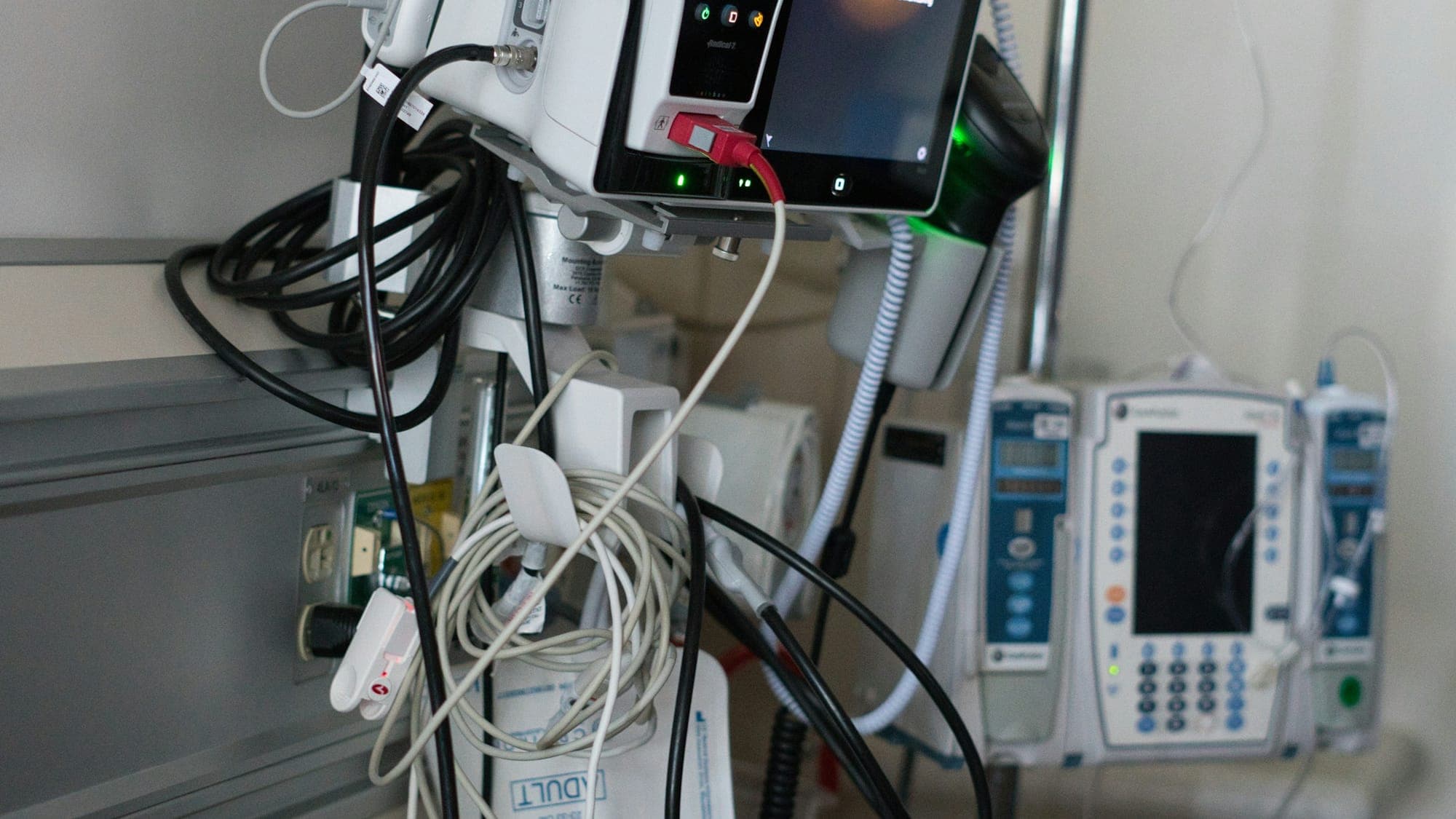Two infusion pumps and a monitor mounted on a bedside rail, their power and infusion lines running down the wall channel behind them
