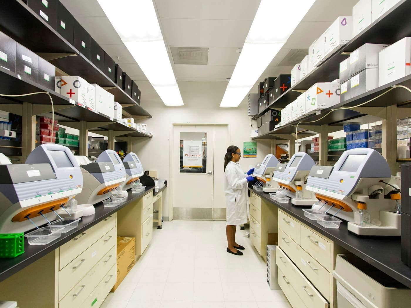 A laboratory aisle with a row of automated bench top analyzers running down both benches, a technician working at the far end