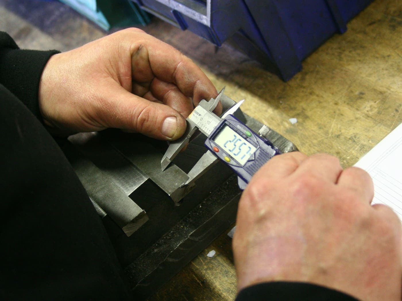 A machined steel block held in one hand and measured with a digital caliper in the other
