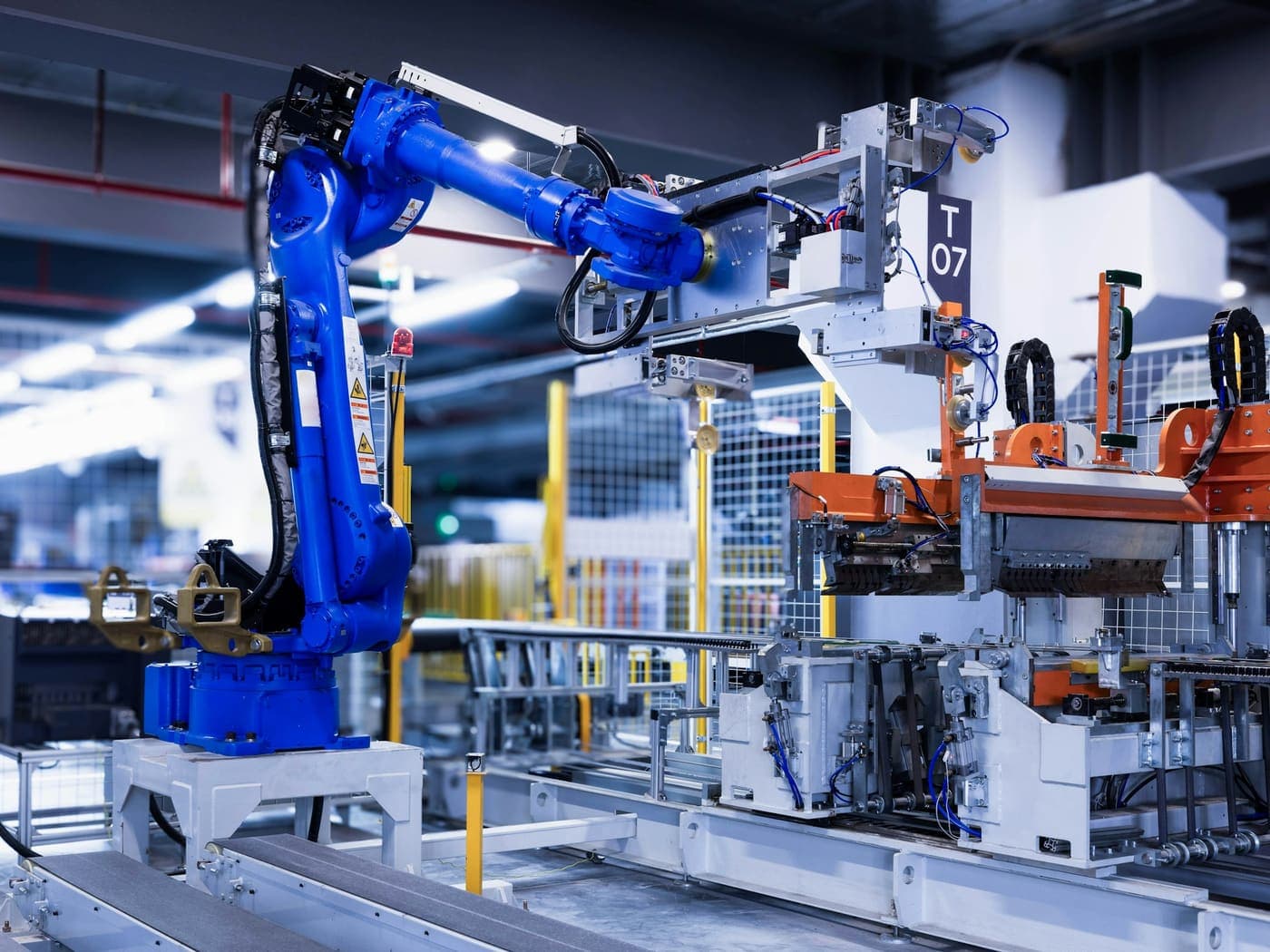 A blue articulated industrial robot on its plinth beside an automated line, its arm reaching over the workpiece carriers