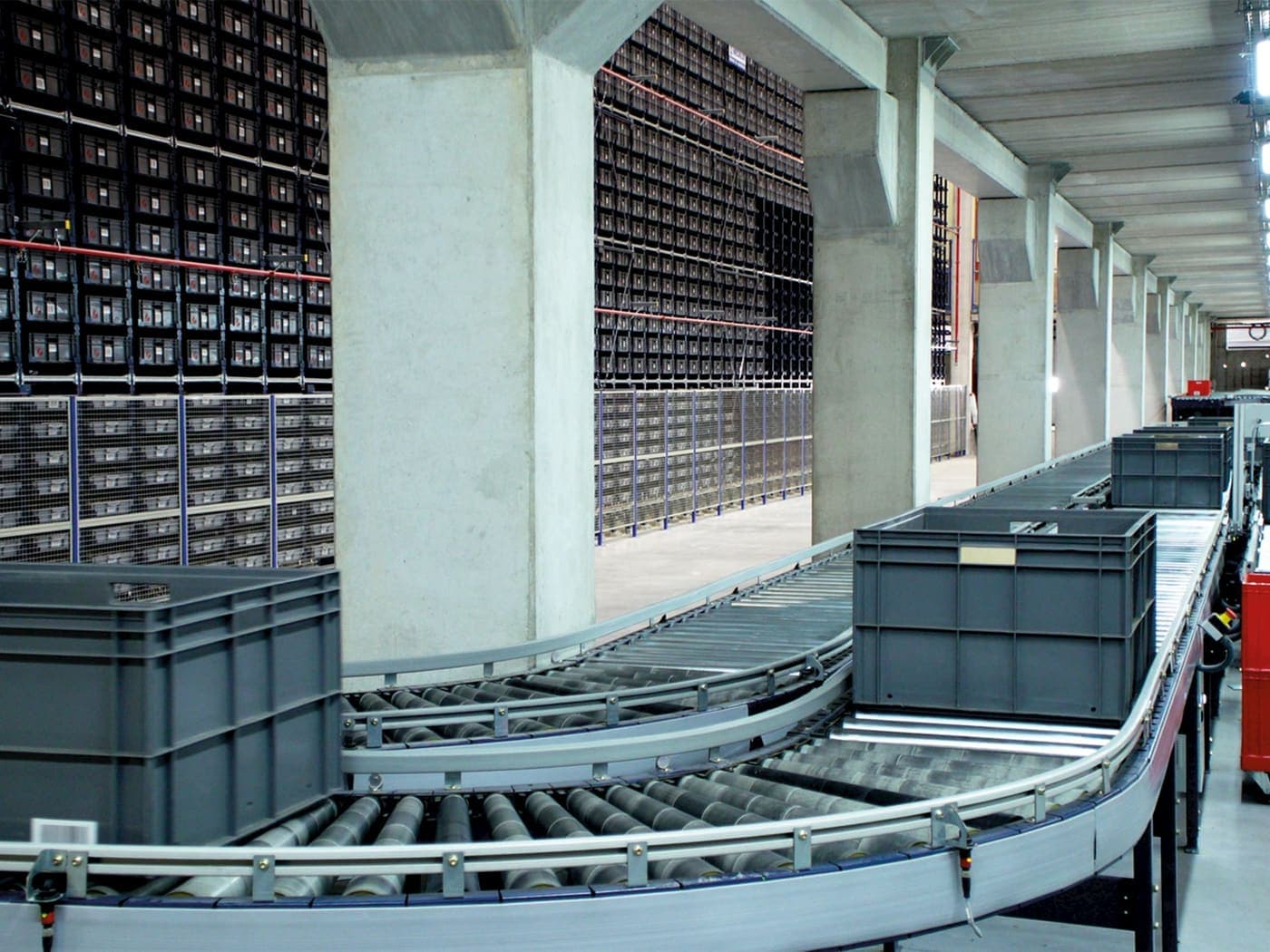 A roller conveyor carrying grey totes past a full height wall of small parts bins in a warehouse