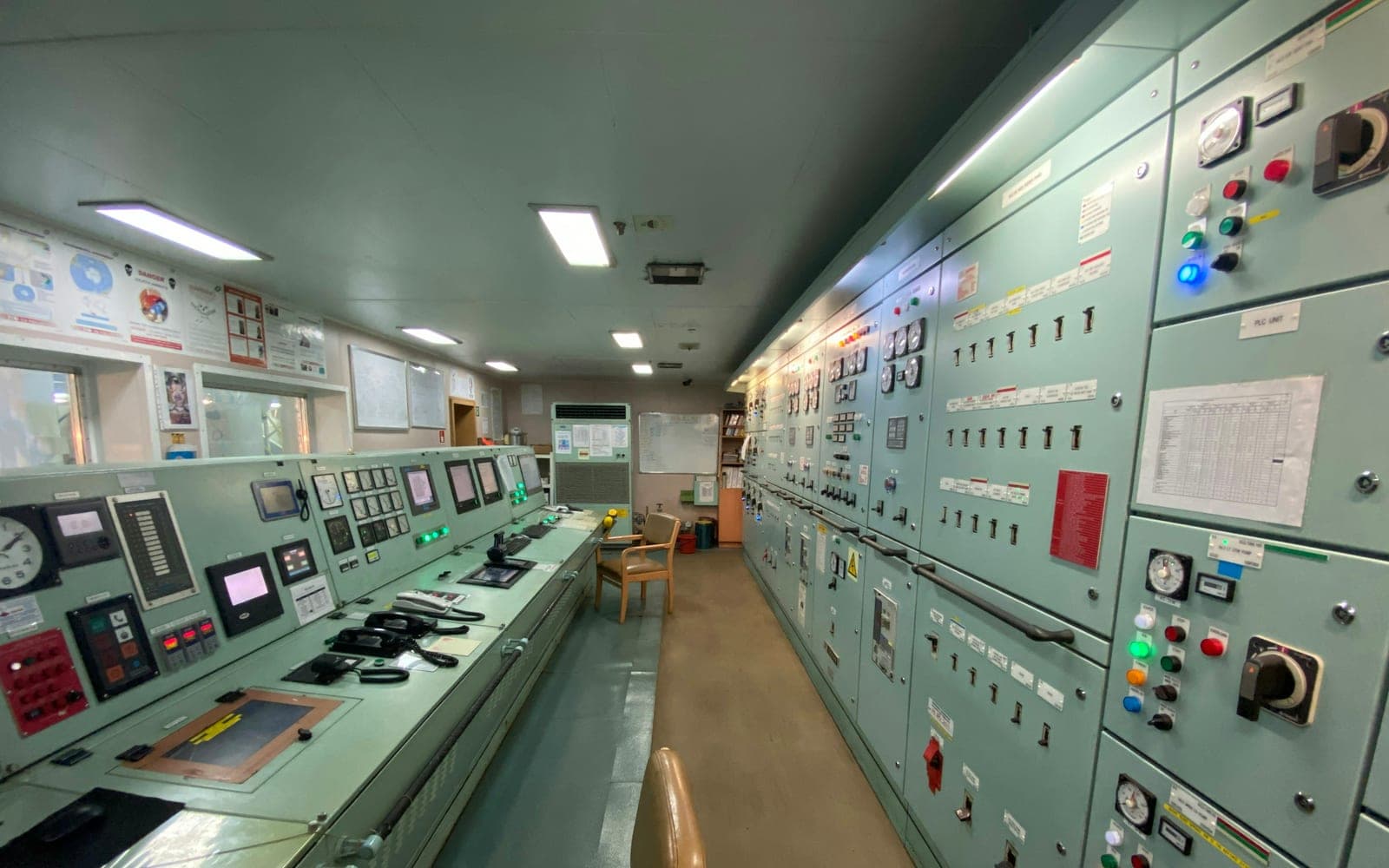 The engine control room of a merchant vessel, its console of alarm panels, gauges and telegraph running down one side and the main switchboard with its breaker cubicles and indicator lamps down the other