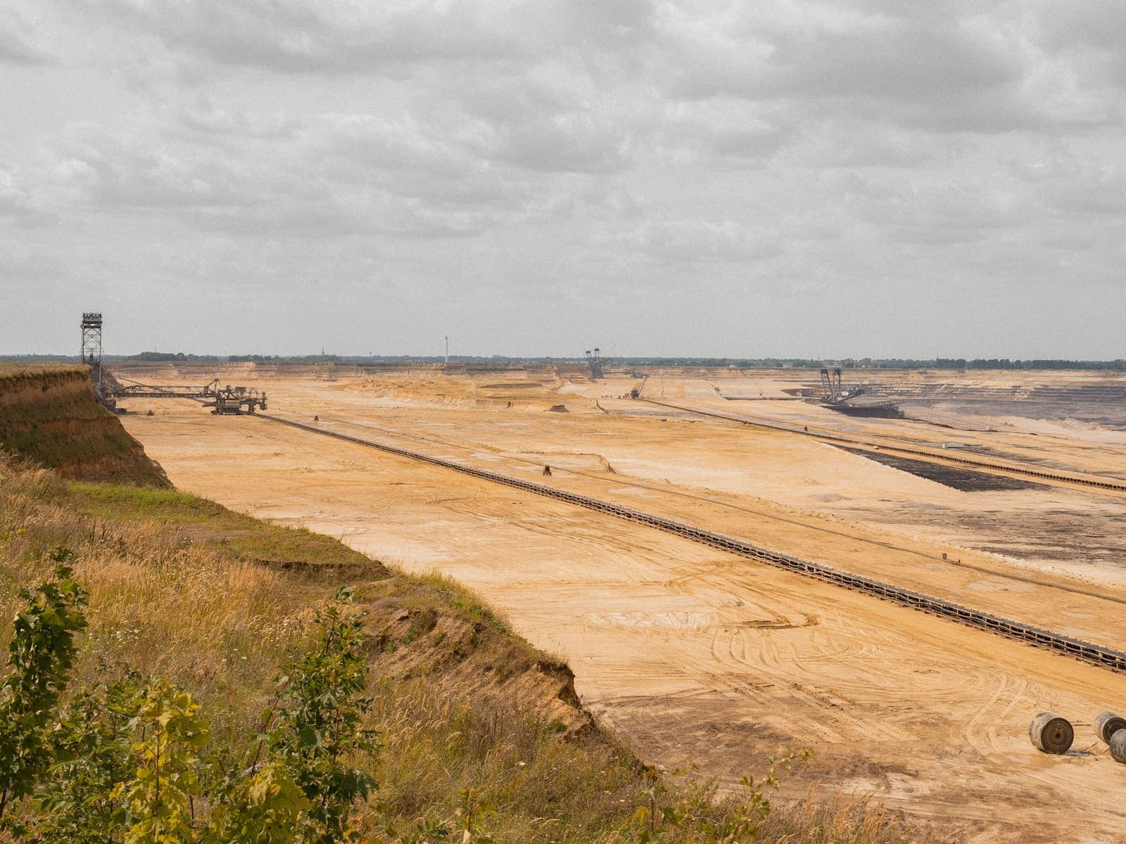 Open cast mine spread to the horizon, with two overland conveyor runs carrying material away from a bucket wheel excavator on the near rim toward machinery kilometres away