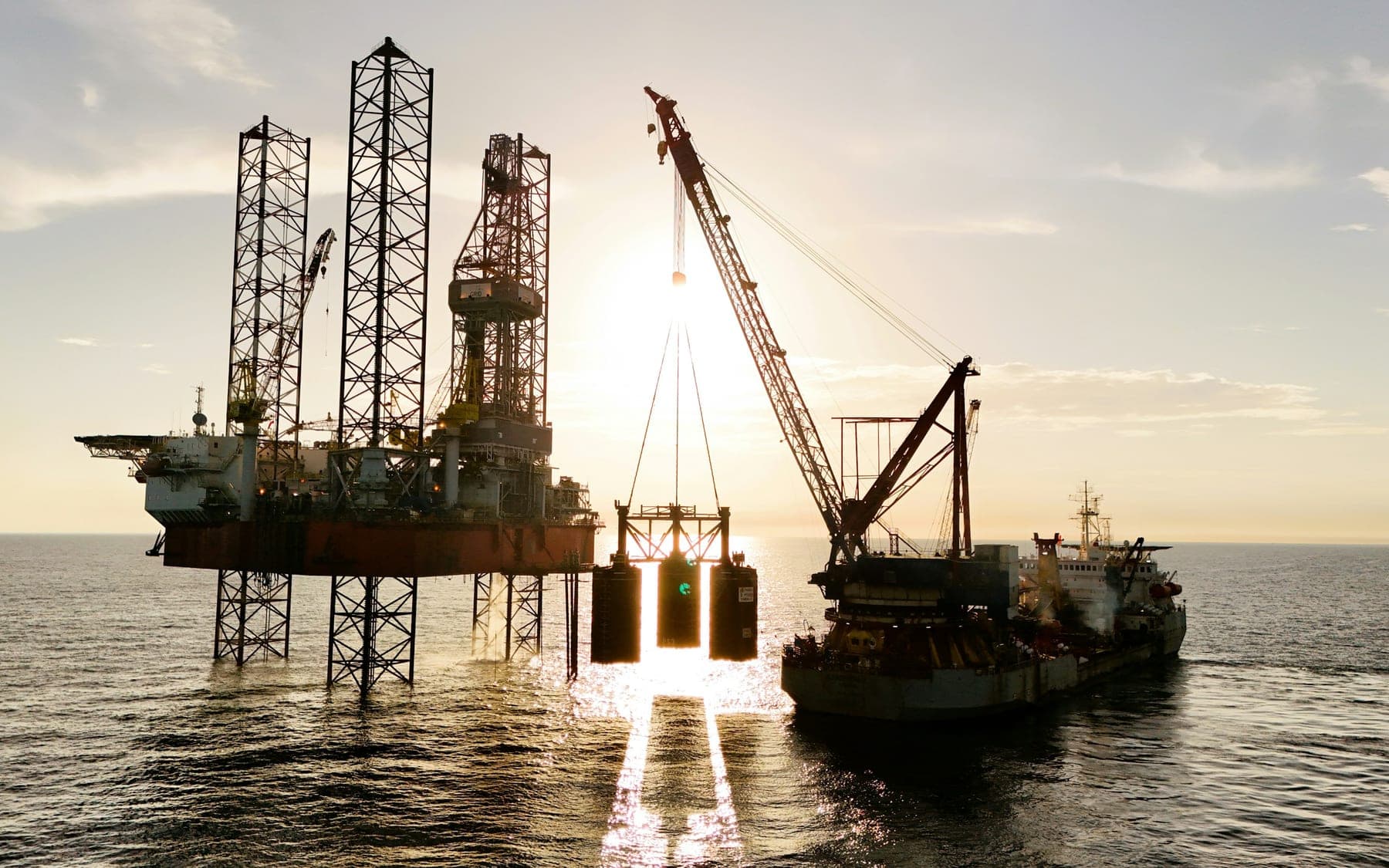 An offshore production platform standing on its legs at sea at sunset, with a crane barge lifting a module alongside it and a support vessel working the same location