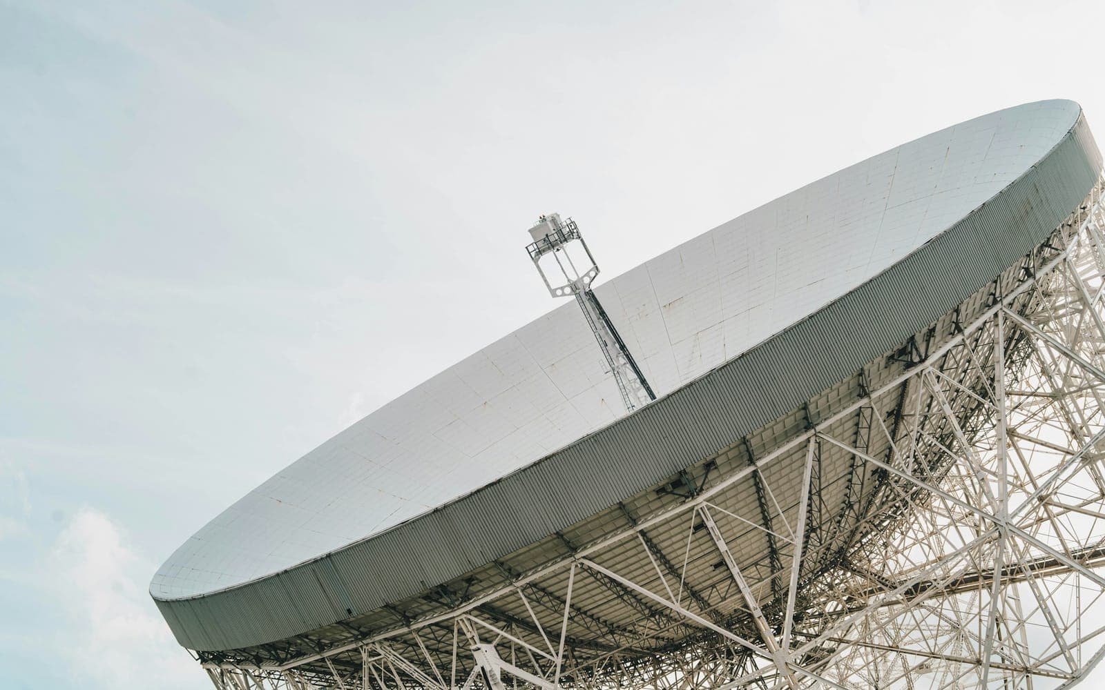A large steerable parabolic antenna seen from beneath its rim, its panelled reflector and lattice backing structure set against a pale sky