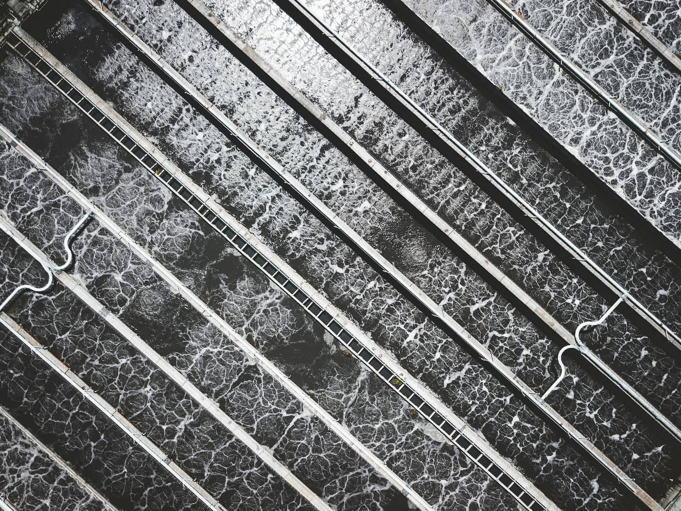Aeration lanes at a wastewater works from above, the surface broken into white foam between concrete dividing walls with a walkway crossing them